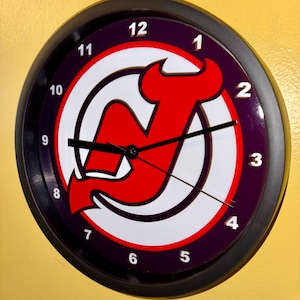 May include: Round wall clock featuring the New Jersey Devils logo in red, white, and black. The clock face has a purple background with white numerals and black hands. The clock is encased in a black frame.