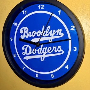 May include: Round wall clock with a black frame and a blue face. The clock face features the words "Brooklyn Dodgers" in white script within a white circle, with black hands and white numerals.