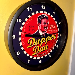 May include: A round black clock with a navy blue border and white numbers. The clock face features a red center with a vintage illustration and the words "Dapper Dan Men's Pomade". The clock also includes the text "Contains Seal Oil" and "Net Wt. 3 oz."