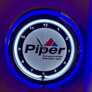 May include: Circular wall clock with a black face, white numerals, and the "Piper" logo. The clock has a blue neon light around the edge and displays the text "THE NEW PIPER AIRCRAFT, INC."
