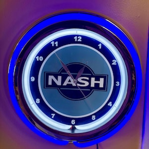 Puede incluir: Un reloj de pared redondo con un borde de luz de neón azul. La esfera del reloj es azul claro con la palabra "NASH" en blanco. El reloj tiene un marco de metal plateado y manecillas negras.