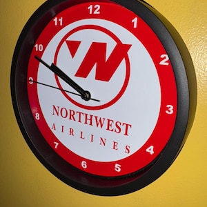 May include: Circular wall clock with a black frame and a red and white face. The clock face displays the Northwest Airlines logo and the words "NORTHWEST AIRLINES". The clock hands are black.