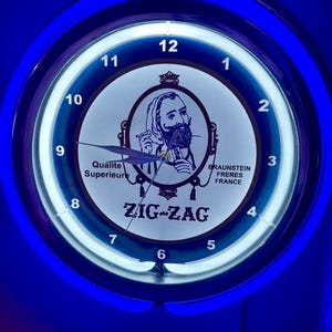 May include: A round wall clock with a bright blue neon light ring. The clock face features the Zig-Zag logo, an illustration of a man, and the text Qualite Superieure and BRAUNSTEIN FRERES FRANCE. It has black clock hands and white numbers 1 through 12.