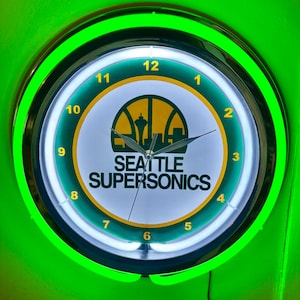 May include: A round wall clock featuring the Seattle SuperSonics logo with a basketball and Space Needle graphic. The clock has a white face, yellow numbers, and a bright green neon light ring around the perimeter. The text SEATTLE SUPERSONICS is printed in black below the central logo.