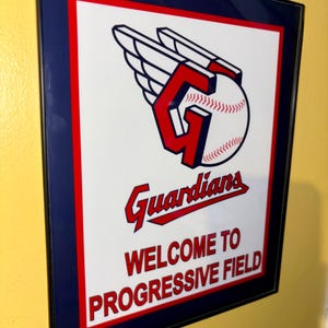 May include: Framed sign featuring the Cleveland Guardians baseball team logo, a stylized "G" with a baseball and wings. The sign reads "Guardians" and "Welcome to Progressive Field" in red text on a white background, framed in red and blue.