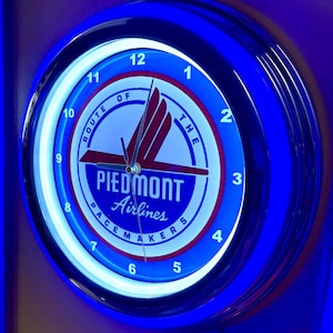 May include: A circular wall clock with a blue and chrome surround. The clock face displays the text "PIEDMONT Airlines PACEMAKERS" in a blue and red design. The clock has a white illuminated border and shows the numbers 1 to 12.