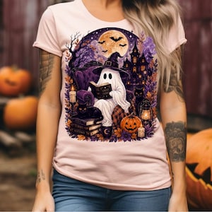 May include: A light pink t-shirt featuring a graphic print of a ghost wearing a witch hat, sitting in a chair and reading a book titled Ghost Stories. The scene includes black cats, a haunted house, a full moon, bats, and a jack-o'-lantern.