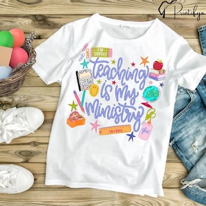 以下が含まれることがあります： 「Teaching is my Ministry」という青い文字が書かれた白いTシャツ。学校をテーマにしたグラフィックで装飾されています。デザインには星、リンゴ、地球儀、学用品が含まれています。木製の表面にTシャツが展示されています。