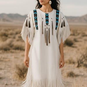 Puede incluir: Vestido color crema con detalles de flecos y abalorios decorativos en negro y turquesa. El vestido presenta mangas cortas y un dobladillo a la rodilla, adecuado para un estilo bohemio o del suroeste.