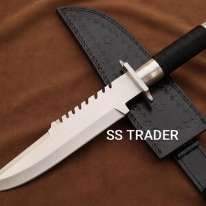 May include: A stainless steel survival knife with a serrated edge and a black wrapped handle. The knife rests on a black leather sheath with the text "SS TRADER".