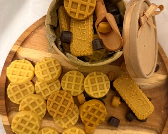 May include: A wooden tray holds an assortment of yellow waffle-shaped and rectangular cookies, along with dark chocolate squares. A round container with a lid and a wooden scoop are also present. The cookies have the word "Lotus" imprinted on them.