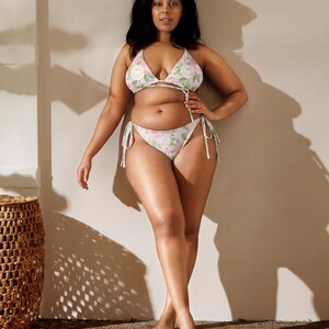 May include: A two-piece bikini set with a floral pattern. The top is a triangle style with adjustable ties, and the bottoms are a classic cut with tie sides. The fabric features a white background with pink and green floral accents. The model is standing on a woven mat.