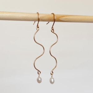 May include: A pair of gold-toned drop earrings featuring a long, slender, wavy metal wire design. Each earring terminates in a small, white, oval-shaped pearl suspended from the bottom curve of the wire, attached via a simple hook mechanism.