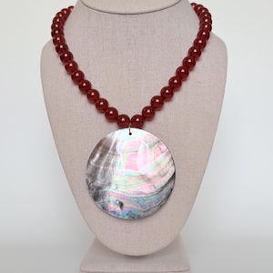 May include: A necklace featuring a large, iridescent pendant and a strand of red beads. The pendant is a large, round shell with a shimmering, multi-coloured surface. The beads are a deep red colour and are strung together to form a necklace.