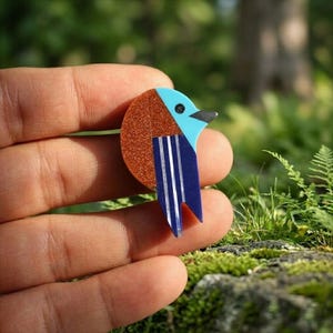 May include: A colorful bird brooch with a geometric design. The brooch features a round, glittery copper-colored body, a light blue head, and a dark blue tail with white stripes. The bird has a black eye and beak. The brooch is held in a hand against a blurred green background.