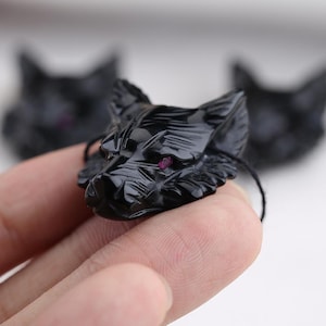 May include: A black carved wolf head pendant with a red gem eye. The pendant is held in a hand, with two other wolf head pendants slightly out of focus in the background. The wolf head is detailed and has a glossy finish.