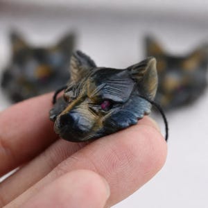 May include: A carved wolf head pendant made of blue and gold tiger eye stone, featuring small, deep pink gemstone eyes and a thin black cord attached to the sides. The wolf carving displays detailed fur texture and pointed ears in a natural stone finish.