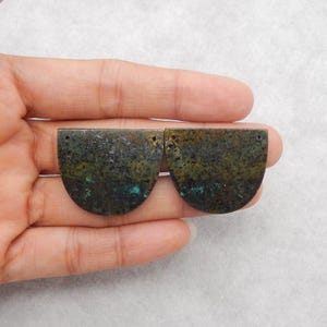 May include: Two semi-circular, dark green and brown stone pendants with turquoise accents. Each pendant has a small hole at the top for stringing. The pendants are held in a hand against a white background.