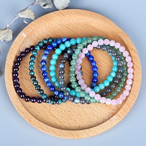 Peut inclure: Un ensemble de huit bracelets en pierres précieuses de différentes couleurs, dont le violet, le bleu, le vert et le rose. Les bracelets sont disposés en cercle sur une assiette en bois.
