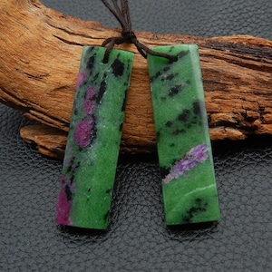 May include: Two rectangular pendants with a green base color, accented with black and magenta spots and streaks. The pendants are strung on a brown cord. The background features a piece of wood and a dark leather-like surface.