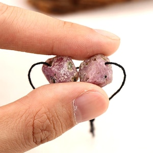 May include: Two rough-cut, pink gemstones with black string loops. The stones are held between fingers against a white background. The gemstones have a natural, unpolished appearance, with variations in color and texture. The stones are likely used for jewelry making.