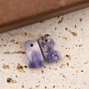 Puede incluir: Dos colgantes rectangulares con esquinas redondeadas, cada uno con un orificio perforado en la parte superior. Un colgante muestra un patrón jaspeado liso en lavanda y blanco, mientras que el otro presenta un diseño más oscuro con vetas moradas.