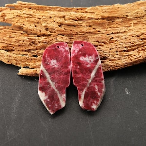 May include: Two flat, irregularly shaped stone pendants featuring a deep red and white marbled pattern. Each piece has a smooth, polished surface and a single small hole drilled near the top edge for suspension, showcasing natural veining.