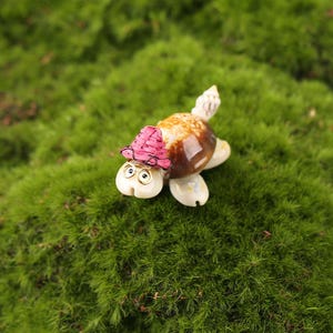 May include: A whimsical miniature turtle figurine with a brown shell, cream-colored limbs, and a pink woven hat. The turtle has gold-rimmed glasses and is set against a backdrop of vibrant green moss.