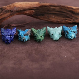 Peut inclure: Cinq pendentifs en forme de tête de loup en pierre sculptée, dans des tons de bleu et de vert. Les pendentifs sont finement sculptés avec des détails précis. Un cordon en cuir marron est attaché à l'un d'eux. Fond marron foncé avec du bois flotté.