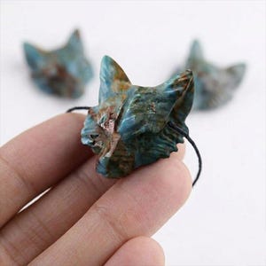 May include: A carved teal and brown stone fox head pendant with a black string. Two other fox head pendants are visible in the background. The fox head is held in a person's hand.