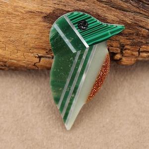 May include: A bird-shaped pendant crafted from green malachite, silver, and a shimmering, speckled material. The bird's head features a striped pattern, and a small, dark circle represents the eye. The pendant is set against a wooden background.