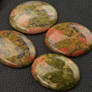 Peut inclure: Quatre pierres d'unakite polies. Les pierres ovales présentent un mélange de teintes vertes, roses et orangées. L'unakite a une finition brillante et est posée sur un fond sombre.