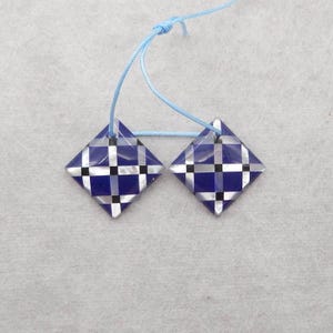 Op de afbeelding: Twee vierkante hangers met een blauw, wit en zwart geblokt patroon. De hangers zijn verbonden door een lichtblauw touwtje. Deze kunnen worden gebruikt voor het maken van sieraden of als oorbellen.