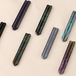 May include: An assortment of gemstone pendants in various colors, including green, blue, and purple. Each pendant has a rectangular shape with a pointed end and two holes at the top. The pendants are arranged on a light-colored surface.