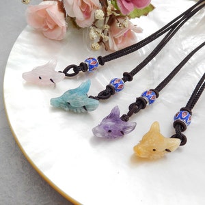 May include: Four gemstone necklaces featuring carved wolf pendants in pink, blue, purple, and yellow. Each pendant is attached to a black cord with a blue and red beaded accent. The necklaces are displayed on a white, pearlescent surface.