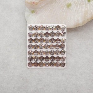 May include: A collection of small, flower-shaped embellishments. Each flower has a dark, iridescent color with a pearlescent sheen. The flowers are arranged in rows on a white card, suitable for crafts or jewelry making.