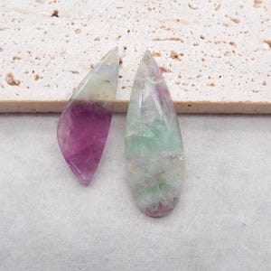 Può includere: Due pietre preziose di fluorite lucidate a forma di goccia. La pietra a sinistra presenta una sfumatura distinta dal viola intenso alla base al verde pallido traslucido in punta. La pietra a destra mostra una miscela screziata di tonalità verde e rosa.