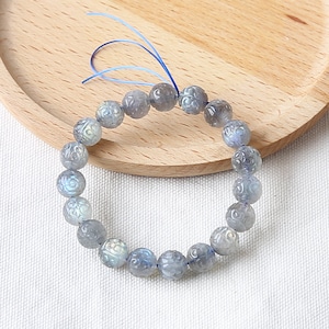 May include: A blue labradorite gemstone bracelet with carved floral designs on each bead. The bracelet is strung on a blue cord.