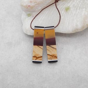 Puede incluir: Dos pendientes rectangulares con un diseño en capas de tonos beige, marrón y amarillo. Cada pendiente tiene detalles en blanco y negro en la parte superior e inferior, unidos por un cordón marrón. Los pendientes están sobre un fondo neutro.