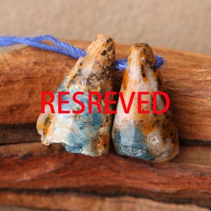 May include: Two cone-shaped beads featuring a mottled pattern of tan, brown, and blue mineral inclusions, strung together on a thin blue thread. The word RESREVED is printed in bold red capital letters across the centre of both stone beads.