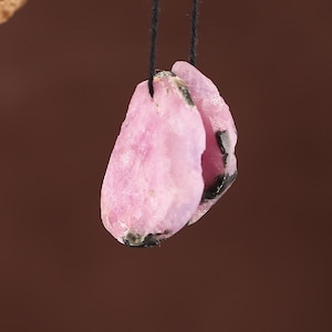 Op de afbeelding: Een hanger met een ruw geslepen, roze edelsteen met donkergrijze insluitsels, hangend aan een zwart koord. De steen heeft een natuurlijke, onregelmatige vorm en een enigszins doorschijnend uiterlijk.