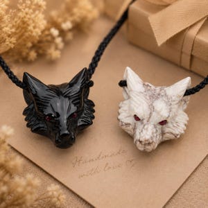 May include: Two wolf head pendants on black braided cords. One pendant is polished black stone, the other is white marbled stone. Both feature small, inset red eyes. The text "Handmade with love" with a heart symbol is printed on the card below.