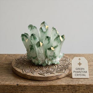 May include: A cluster of green phantom crystals with gold accents, displayed on a carved wooden coaster. A tag reads "Green Phantom Crystal". The crystals are a light green color and have a natural, organic shape.