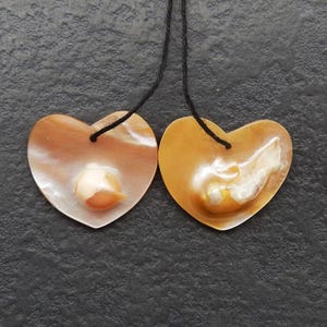 May include: Two heart-shaped pendants with a pearlescent finish, each suspended by a black cord. The pendants have a warm, golden-brown hue with iridescent highlights and a small, off-white accent in the center.