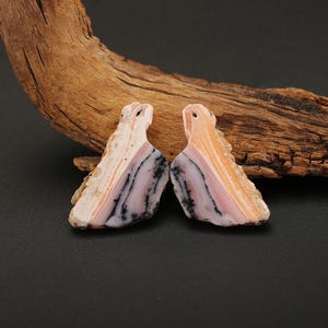 May include: Two natural stone pendants with a unique shape and color pattern. The pendants feature a mix of pink, white, and black stripes, with a hole at the top for stringing. The background includes a piece of driftwood.