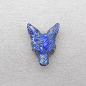Peut inclure: Une tête de loup sculptée en lapis-lazuli. La sculpture présente une gravure détaillée du visage d'un loup, avec des oreilles pointues et une surface texturée. La pierre est d'un bleu profond avec des veines blanches et grises.