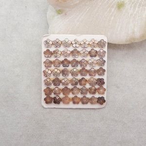 May include: A rectangular card with rows of small, flower-shaped beads in shades of brown and tan. The beads are arranged in a grid pattern, with each flower having five petals. The card is on a light gray surface.