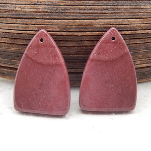 Puede incluir: Dos colgantes triangulares de piedra preciosa de color rosa rojizo. Cada colgante tiene un pequeño agujero en la parte superior para ensartar. Las piedras preciosas tienen una textura moteada y una banda más clara en el medio. El fondo es una superficie de madera.