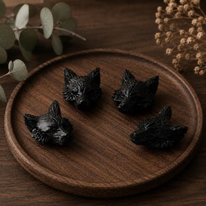 Könnte beinhalten: Vier kleine, schwarze, aus Stein geschnitzte Wolfskopffiguren, die auf einem runden Holztablett angeordnet sind. Jede Figur weist detaillierte Schnitzereien auf, darunter spitze Ohren, eine Schnauze und ein strukturiertes Fellmuster.