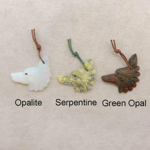 Puede incluir: Tres colgantes de piedra tallada con forma de cabeza de lobo. Los colgantes son de opalita, serpentina y ópalo verde. Cada uno tiene un cordón anudado para colgar. El texto debajo dice "Opalite", "Serpentine" y "Green Opal".